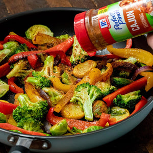 A hand sprinkles McCormick Perfect Pinch Cajun Seasoning on a pan of roasted vegetables.