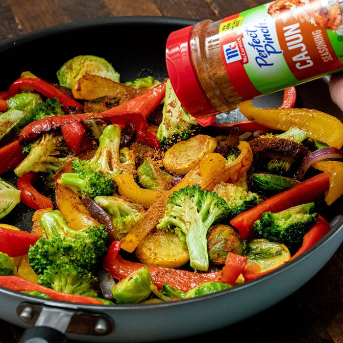 A hand sprinkles McCormick Perfect Pinch Cajun Seasoning on a pan of roasted vegetables.
