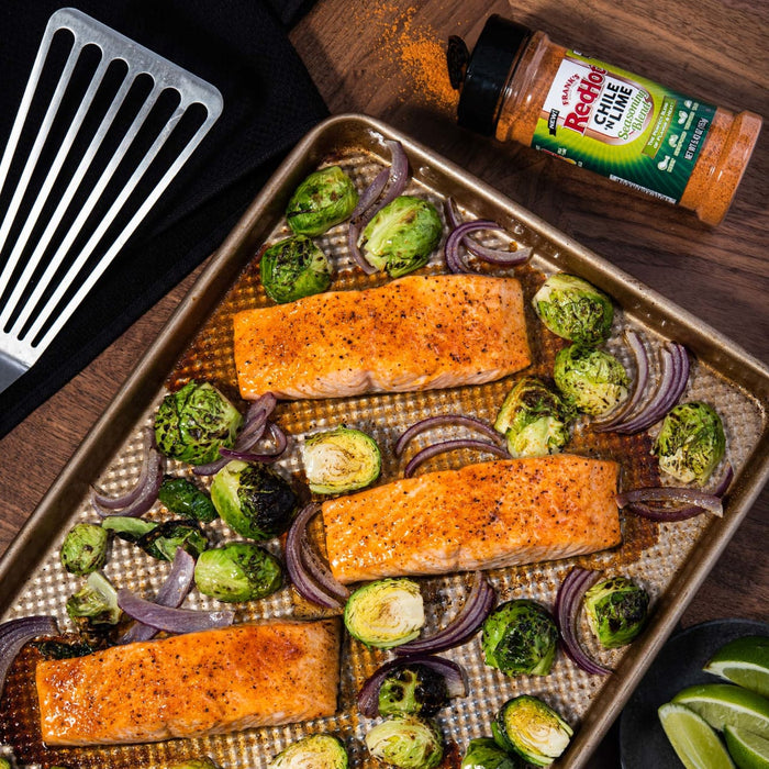 A baking sheet with three salmon fillets, brussel sprouts, red onion, and Franks RedHot Chili n Lime seasoning.