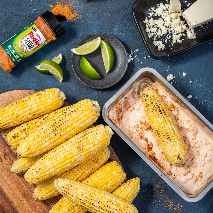 Grilled corn on the cob, lime wedges, and a container of RedHot Chili n Lime seasoning blend.