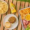 A bottle of French'S Minion Yellow Mustard, 8 Oz sits on a picnic table next to a hamburger topped with mustard and a snack tray with crackers, cheese, and salami.