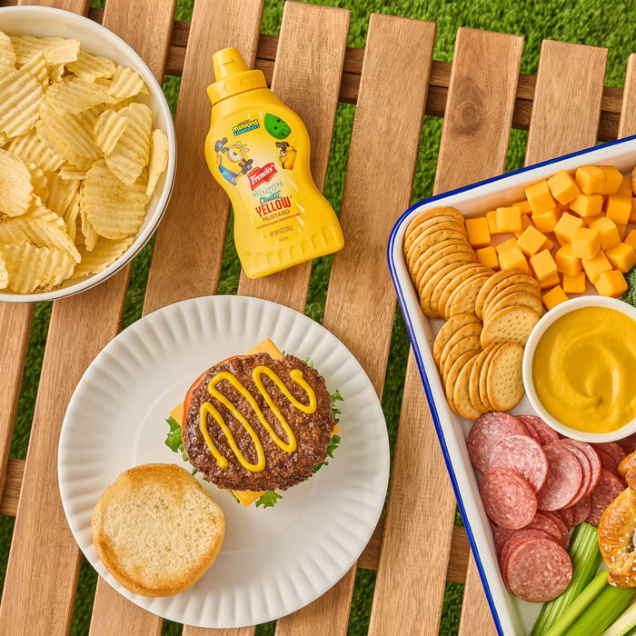 A bottle of French'S Minion Yellow Mustard, 8 Oz sits on a picnic table next to a hamburger topped with mustard and a snack tray with crackers, cheese, and salami.