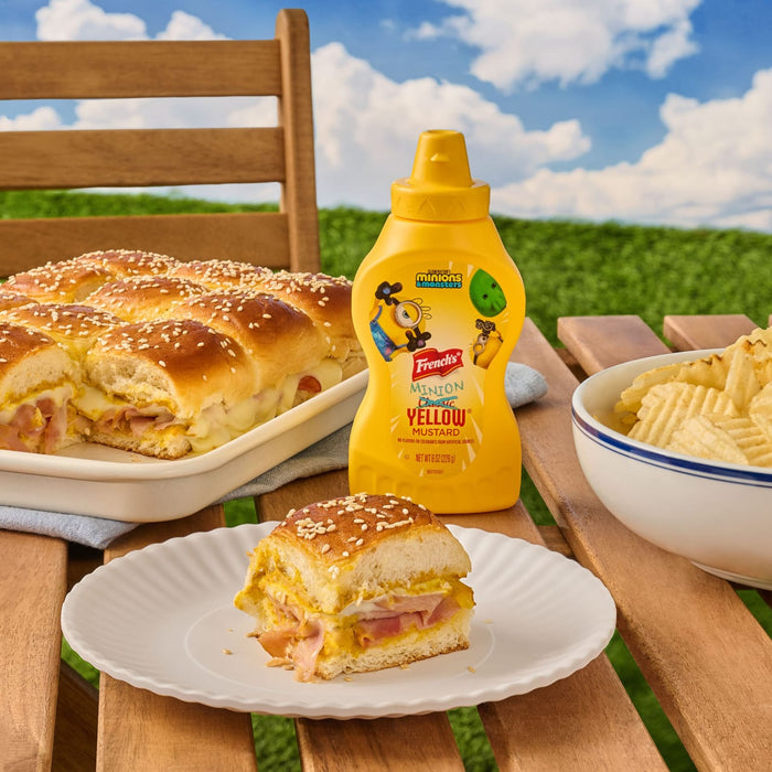 A bottle of French'S Minion Yellow Mustard, 8 Oz is featured in an outdoor picnic setting next to a tray of ham and cheese sliders and a bowl of chips.