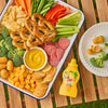 A bottle of French'S Minion Yellow Mustard, 8 Oz, sits next to a large snack platter featuring pretzels, cheese, and vegetables, with the mustard also served as a dip for mini corn dogs.