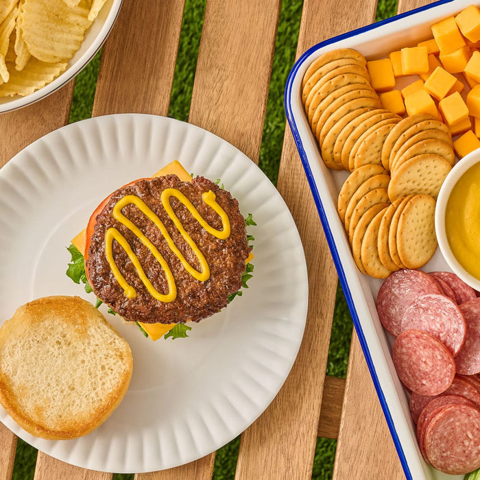 An overhead shot shows a picnic spread with a hamburger on a paper plate, topped with a zigzag of French'S Minion Yellow Mustard, 8 Oz, alongside a tray of crackers, cheese, and salami.