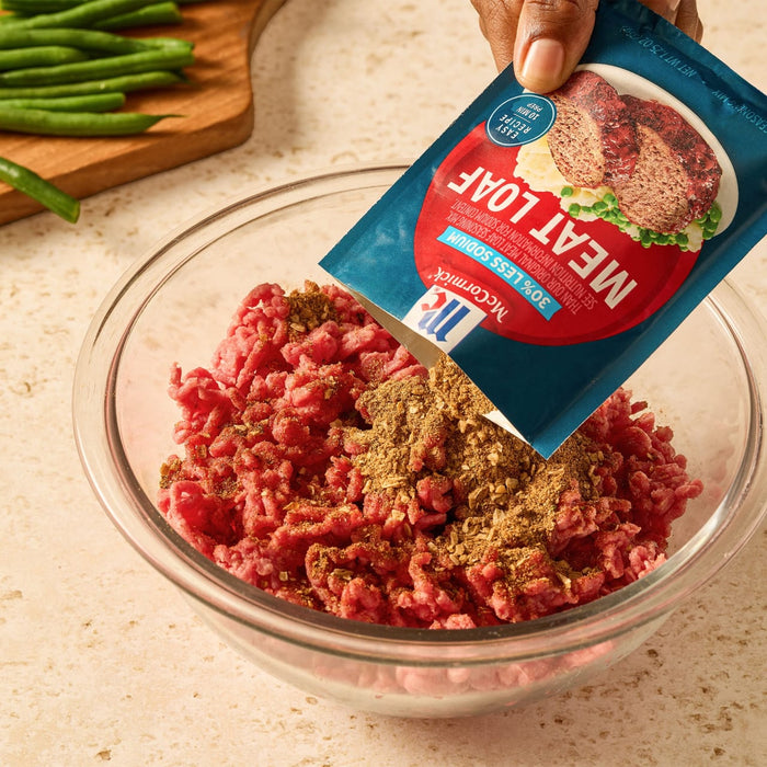 A person pours a packet of Mccormick Regular Meat Loaf Seasoning Mix 1.25 Oz into a glass bowl of raw ground beef to prepare a meal.