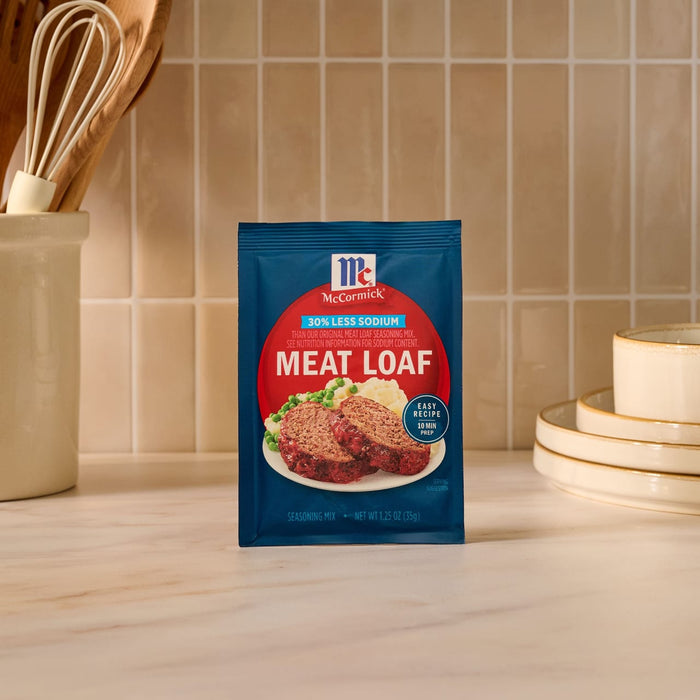 A blue packet of Mccormick Regular Meat Loaf Seasoning Mix 1.25 Oz is displayed on a kitchen countertop, with a utensil holder and a stack of plates in the background.