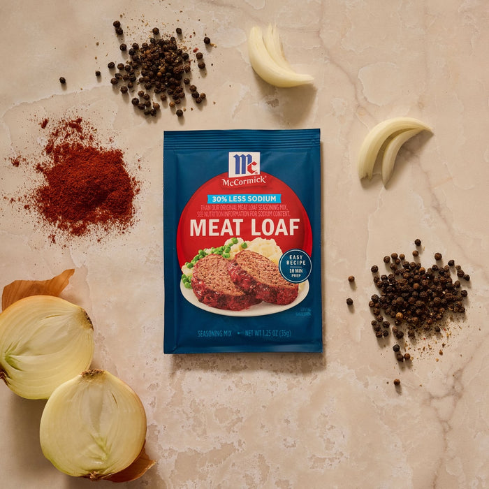 A packet of Mccormick Regular Meat Loaf Seasoning Mix 1.25 Oz is displayed on a countertop surrounded by fresh ingredients like onion, peppercorns, and paprika.