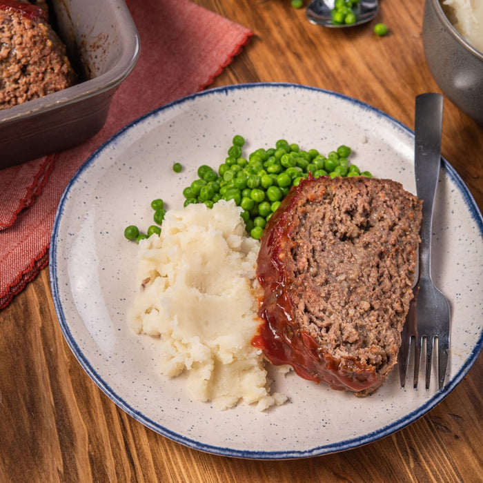 A slice of savory meatloaf, prepared using Mccormick Regular Meat Loaf Seasoning Mix 1.25 Oz, is plated with creamy mashed potatoes and bright green peas for a classic comfort meal.