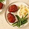 A delicious meal of individual meatloaves, made with Mccormick Regular Meat Loaf Seasoning Mix 1.25 Oz, is served on a white plate with mashed potatoes and green beans.