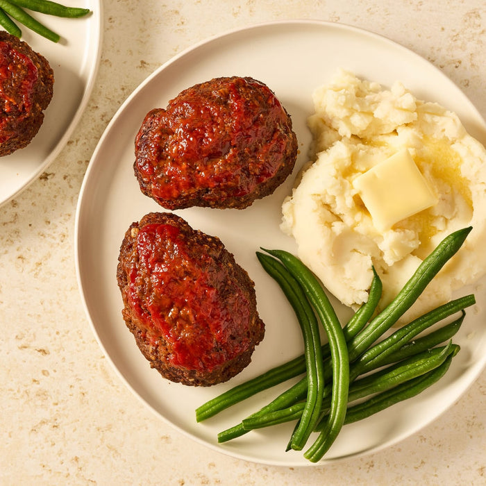 A delicious meal of individual meatloaves, made with Mccormick Regular Meat Loaf Seasoning Mix 1.25 Oz, is served on a white plate with mashed potatoes and green beans.