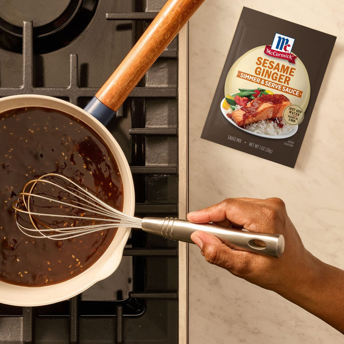 A hand whisks a sesame ginger sauce in a pan on a stovetop, with a package of the Mccormick Sesame Ginger All Purpose Sauce Mix Envelope 1 Oz displayed on the counter.