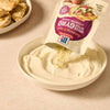 To prepare a tzatziki sauce, a hand pours the contents from a packet of Mccormick Regular Sauce And Seasoning Mix Envelope 1.5 Oz, labeled as Greek Style Gyro, into a bowl of creamy Greek yogurt.