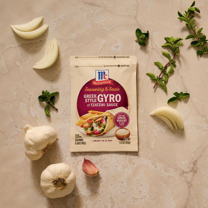 A packet of Mccormick Regular Sauce And Seasoning Mix Envelope 1.5 Oz for Greek Style Gyro is displayed on a countertop surrounded by fresh ingredients like garlic and oregano.
