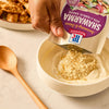 A person prepares a creamy garlic sauce by pouring the Mccormick Regular Sauce And Seasoning Mix Envelope 1.37 Oz into a white bowl of Greek yogurt, with a plate of cooked shawarma chicken in the background.