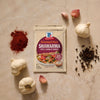 A flat lay product shot shows a Mccormick Regular Sauce And Seasoning Mix Envelope 1.37 Oz for Shawarma, surrounded by ingredients like garlic, paprika, and black peppercorns.
