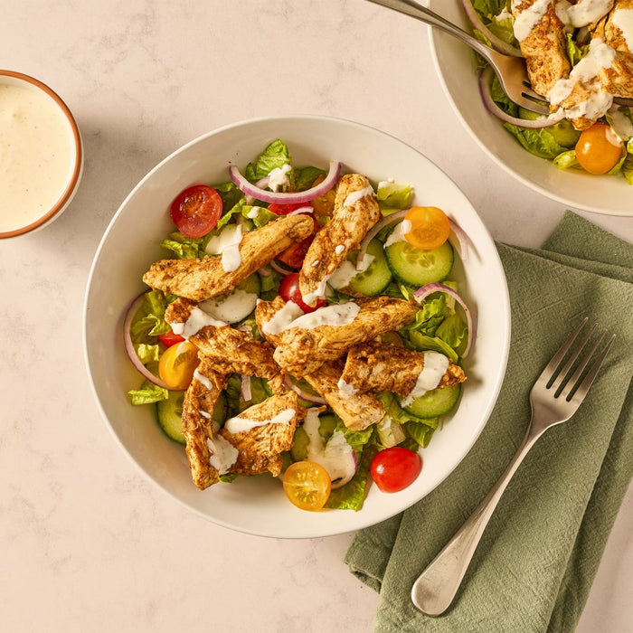 A fresh chicken salad with lettuce, tomatoes, and a creamy dressing is shown in a white bowl, with the chicken seasoned and the dressing prepared using Mccormick Regular Sauce And Seasoning Mix Envelope 1.37 Oz.