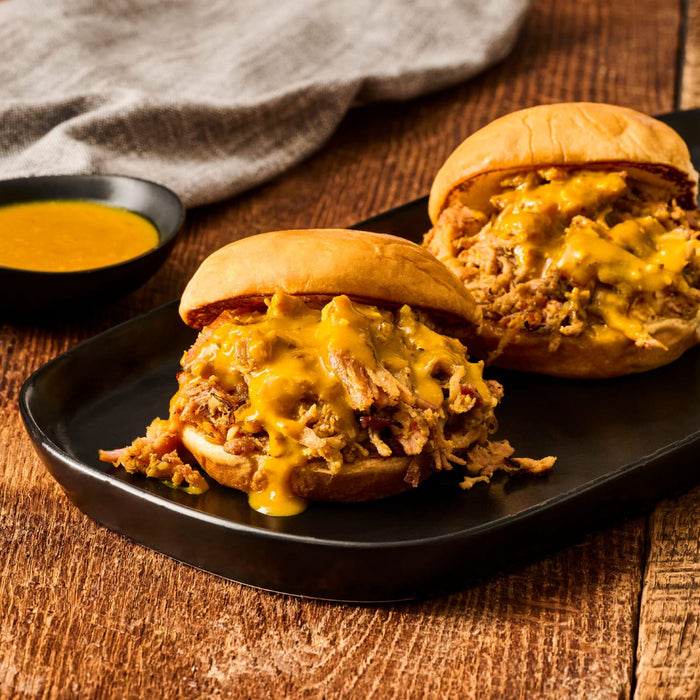 Two pulled pork sandwiches with a yellow sauce are displayed on a black plate, accompanied by a small bowl of the same sauce.