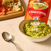 A hand pours a packet of Mccormick Regular Seasoning Mix 1 Oz into a bowl of mashed avocado to make fresh guacamole, with chopped tomatoes and onions on a cutting board in the background.