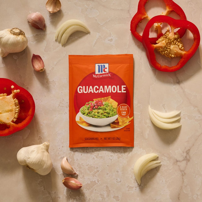 A packet of Mccormick Regular Seasoning Mix 1 Oz is displayed on a countertop surrounded by fresh ingredients like garlic, onion, and red bell pepper.