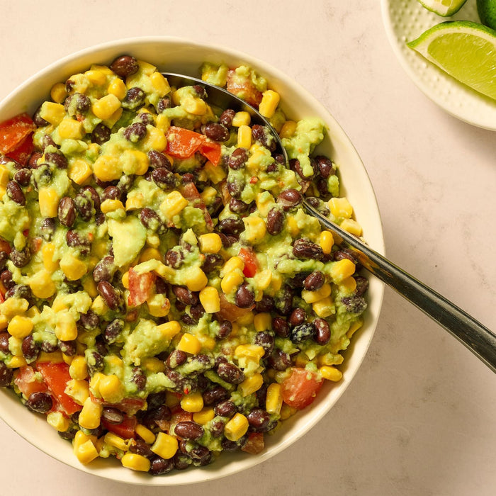 A white bowl filled with a vibrant black bean, corn, and avocado dip, made using Mccormick Regular Seasoning Mix 1 Oz, is shown from above with a serving spoon and lime wedges on the side.