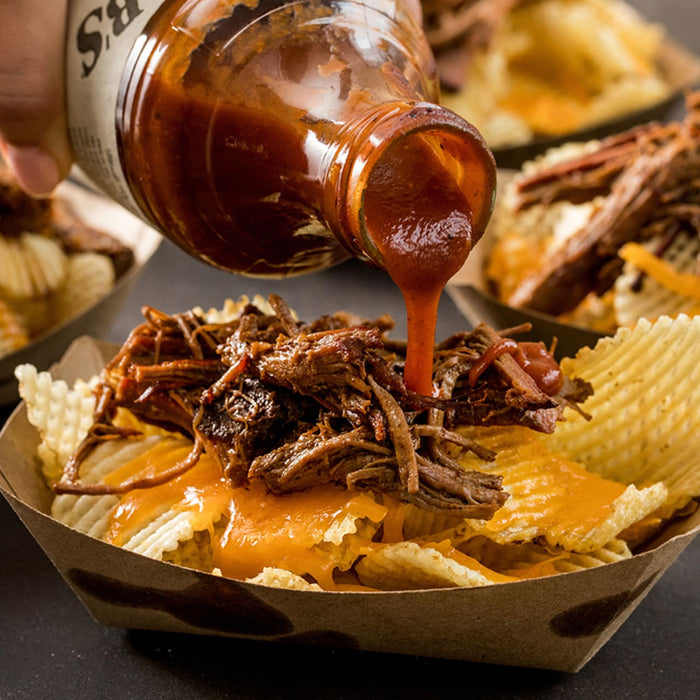 A bottle of S.R. sauce is being poured over a bowl of potato chips shredded beef and melted cheese.