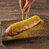 A hand sprinkles seasoning on a piece of meat on a wooden cutting board.