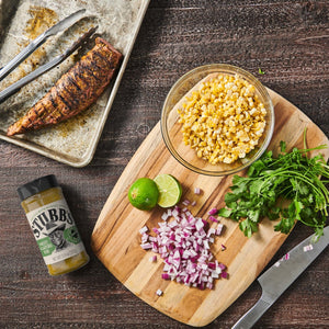 A wooden cutting board with ingredients for a recipe, including corn, red onion, cilantro, limes, and a bottle of Stubbs Sweet Jalapeno Rub.
