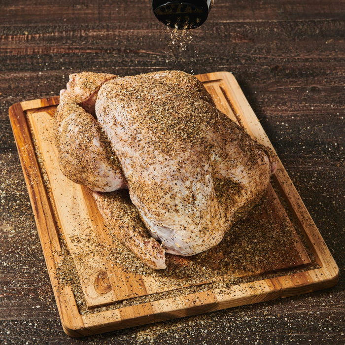 A raw chicken is seasoned with salt and pepper on a wooden cutting board.