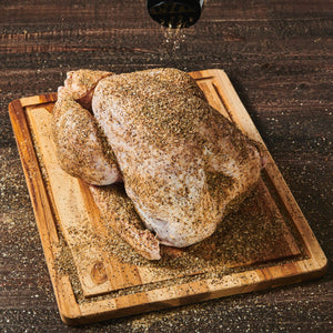 A raw chicken is seasoned with salt and pepper on a wooden cutting board.