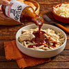 A bowl of shredded chicken is being drizzled with Stubb's BBQ sauce, with coleslaw and buns in the background.