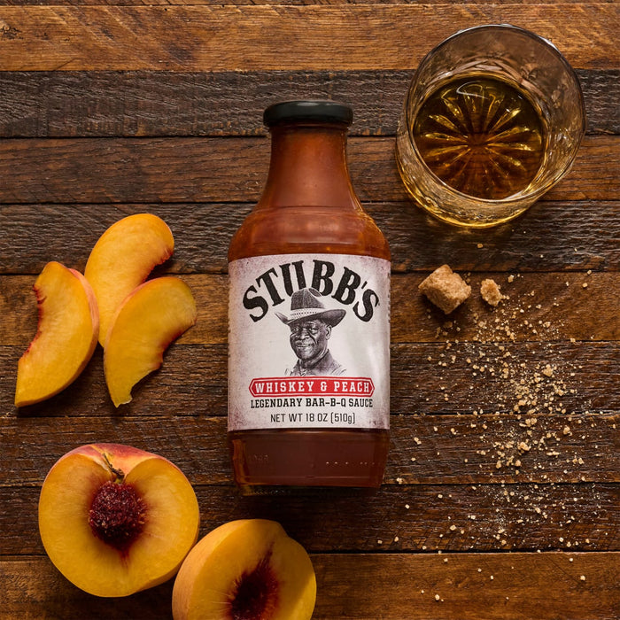 A bottle of Stubb's Sweet Heat Barbecue Sauce is displayed with fresh peaches, brown sugar, and a glass of whiskey on a wooden surface.