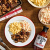 A plate of Stubb's barbecue with mac and cheese and coleslaw is displayed on a wooden table alongside a bottle of Stubb's barbecue sauce.