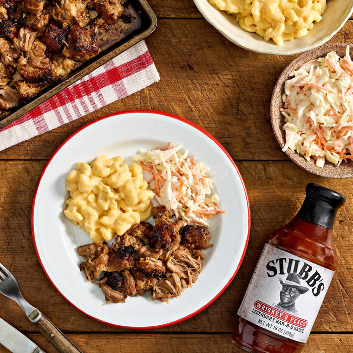 A plate of Stubb's barbecue with mac and cheese and coleslaw is displayed on a wooden table alongside a bottle of Stubb's barbecue sauce.