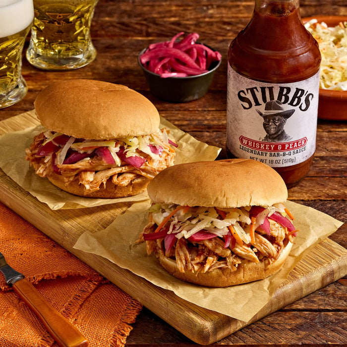 Two Stubb's BBQ sandwiches topped with coleslaw and pickled onions are displayed on a wooden board with a bottle of Stubb's BBQ sauce in the background.