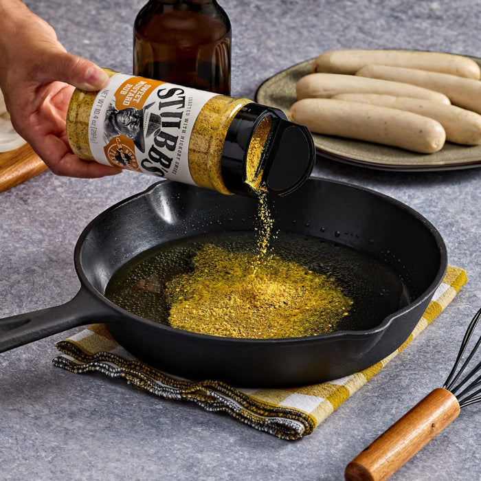 A hand sprinkles Stubbs seasoning into a cast iron skillet with oil, preparing to cook sausages.
