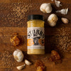 A bottle of Stubb's Honey Pecan BBQ Rub is displayed on a wooden surface with garlic bulbs, honeycombs, and scattered spices.