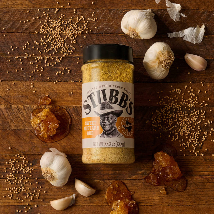 A bottle of Stubb's Honey Pecan BBQ Rub is displayed on a wooden surface with garlic bulbs, honeycombs, and scattered spices.