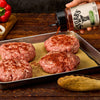 A hand sprinkles Stubbs Grlc Lime E seasoning onto four raw ground meat patties arranged on a baking sheet lined with parchment paper.
