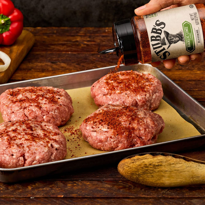 A hand sprinkles Stubbs Grlc Lime E seasoning onto four raw ground meat patties arranged on a baking sheet lined with parchment paper.