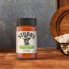 A bottle of Stubbs Grlc Lime E seasoning sits on a rustic wooden surface next to a basket of fresh garlic bulbs against a textured blue background.