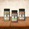 Three bottles of Stubb's seasoning including Hickory Mustard, Stubbs Grlc Lime E, and Texas Smokehouse Style are displayed on a wooden surface against a textured wall.