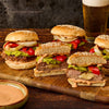 A wooden board holds several juicy burgers stuffed with cheese and topped with guacamole and grilled vegetables seasoned with Stubbs Grlc Lime E alongside a cold beer.