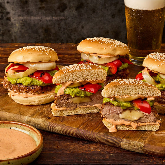 A wooden board holds several juicy burgers stuffed with cheese and topped with guacamole and grilled vegetables seasoned with Stubbs Grlc Lime E alongside a cold beer.
