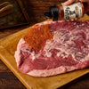 A hand sprinkles Stubbs Smkhouse E seasoning onto a large raw brisket resting on a wooden cutting board.