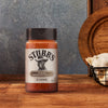 A bottle of Stubbs Smkhouse E seasoning sits on a rustic wooden table next to a basket of fresh garlic bulbs.