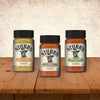 Three bottles of Stubb's seasoning including Hickory Mustard, Garlic Lime, and the Stubbs Smkhouse E variety are displayed on a rustic wooden table against a textured wall.