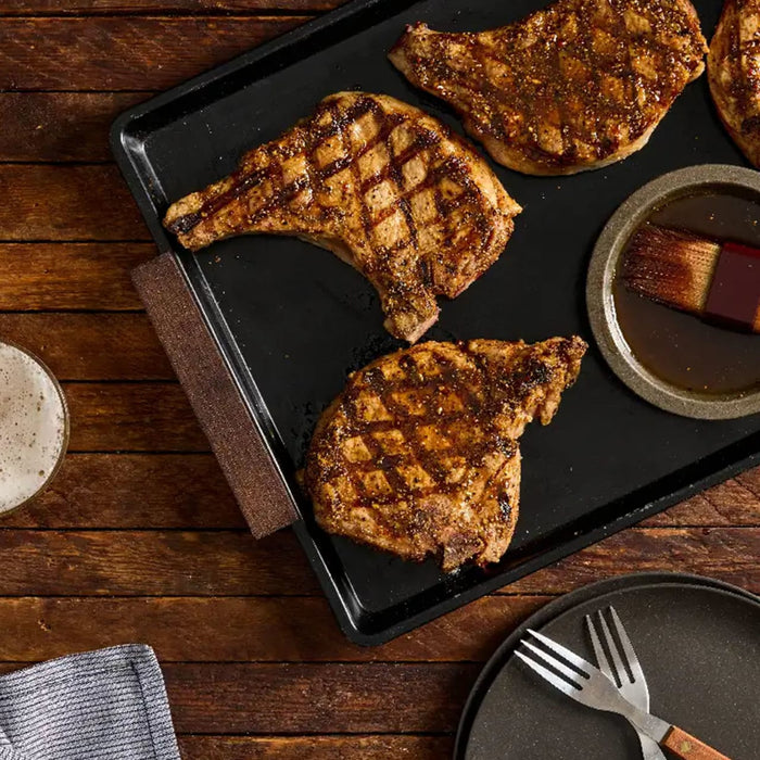 Three bone-in pork chops with cross-hatched grill marks rest on a black griddle pan after being seasoned with Stubbs Smkhouse E.