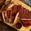 A wooden cutting board holds a rack of sliced ribs seasoned with Stubbs Smkhouse E and served with a side of barbecue sauce.