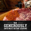 A hand shakes a bottle of Stubbs Smkhouse E seasoning to rub generously onto a large piece of raw meat before cooking.